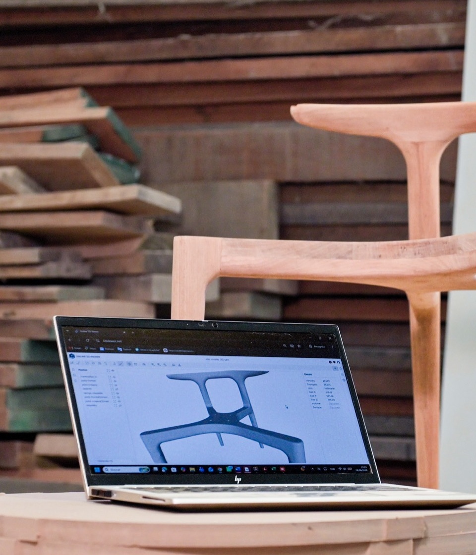 Portátil con el modelo 3D de una silla junto a la silla de madera ya fabricada en el taller